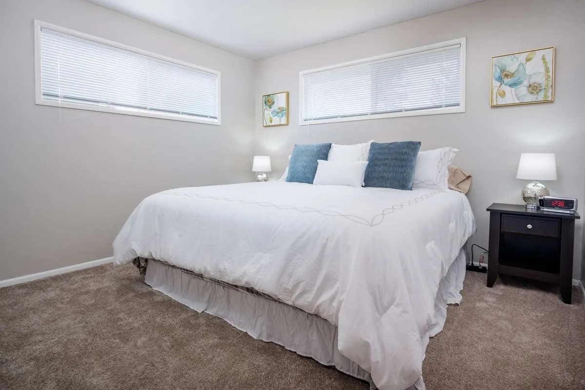 A tranquil king-size bedroom featuring a plush bed with fresh linens, soft lighting from bedside lamps, decorative artwork above the headboard, and natural sunlight streaming through the windows. The room’s calming color palette and thoughtful decor create a peaceful ambiance ideal for restful nights.