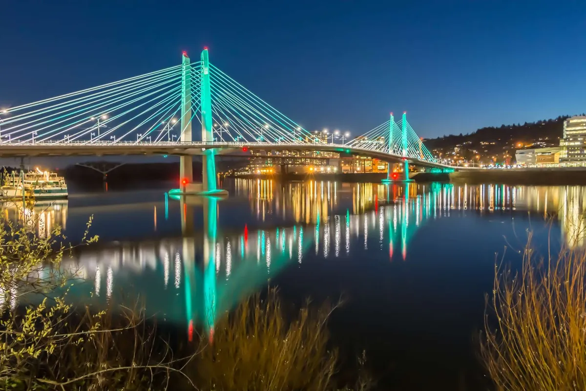 An illuminated bridge stretches over tranquil water, with reflections from city lights creating a lively yet contemplative evening scene. The backdrop of vibrant city life hints at nearby restaurants, evening strolls, and waterfront exploration.