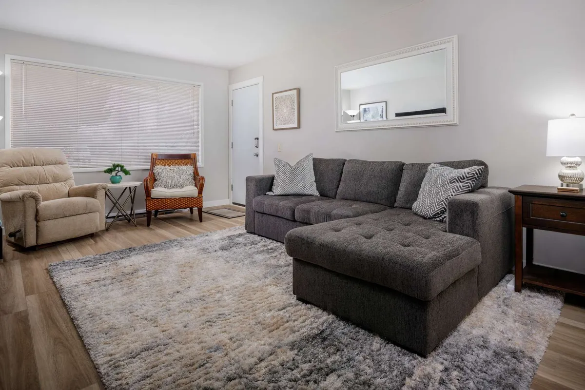 A bright and inviting living room featuring a plush sectional sofa, armchair, coffee table, soft area rug, and stylish mirror. The arrangement is ideal for relaxing, reading, socializing, or enjoying a movie night, with decor that exudes a contemporary, cozy vibe.