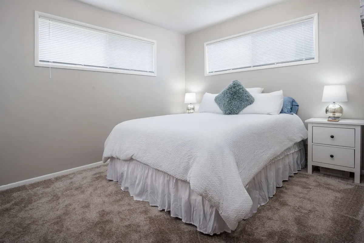 A tranquil bedroom featuring a neatly made bed with decorative pillows, a nightstand with an elegant table lamp, and soft natural light filtering into the space. The calm décor and plush bedding create an inviting atmosphere ideal for relaxation and restful mornings.