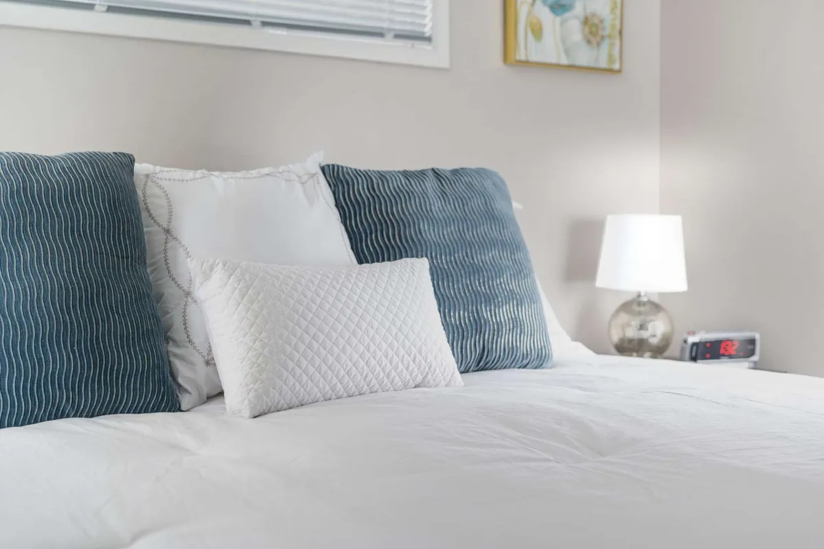 A cozy queen bedroom scene with blue striped and white quilted pillows, a bedside lamp emitting a soft glow, and an alarm clock on the nightstand. The room is bathed in gentle natural light, enhancing the inviting and restful setting for guests.